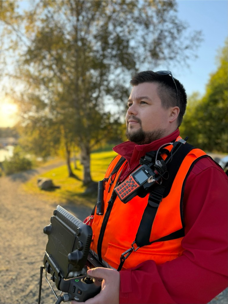 Portrettfoto av dronepilot med refleksvest og radio under flyving i solnedgang